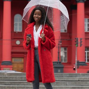 Woman wearing a large red coat, jeans, and sneakers is holding a transparent umbrella in one hand and a coffee mug in the other.