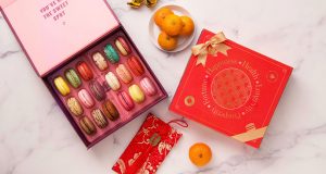  A box full of 18 assorted macarons has a Woops! Macaron box with a red Lunar New Year sleeve to the right. Lying around are some red envelopes and tangerines