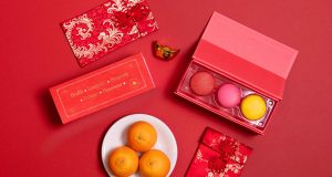 A box full of three assorted macarons has a Woops! Macaron box with a red Lunar New Year sleeve to the left. Surrounding them are some red envelopes and a plate full of tangerines. 