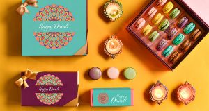 A box full of 18 assorted macarons is surrounded by macaron boxes with Diwali sleeves, assorted macarons, and Diwali candles.