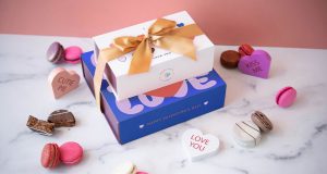: A stack of two French macaron boxes with Valentine’s Day sleeves surrounded by heart-shaped candies, macarons, and alfajores