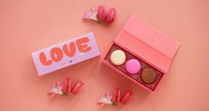  A box full of three French macarons has a macaron box with a red and pink Valentine’s Day sleeve to the left. Above and below are some French macarons and flowers