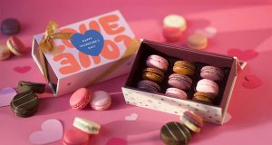A box full of nine French macarons has a macaron box with a Valentine’s Day sleeve to the left. Surrounding them are some assorted alfajores, macarons, a paper hearts. 