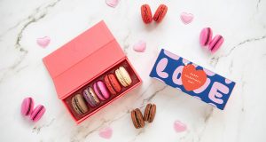 A box full of five French macarons has a macaron box with a blue Valentine’s Day sleeve to the left. Lying around are some French macarons and tiny pink hearts.