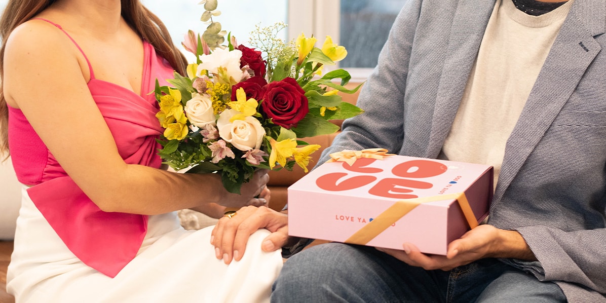 A smiling couple is holding a Woops! French macaron box with a Valentine’s Day sleeve and some flowers between them.