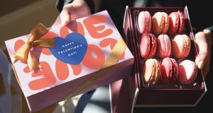 A box full of assorted macarons and a macaron box with a Valentine’s Day sleeve are being held by a pair of hands. 