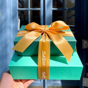 A stack of two green French macaron boxes tied together by a golden ribbon with Woops! Inscriptions