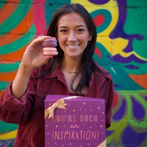 A smiling woman is holding a box of macarons with a purple sleeve and two French macarons in her other hand. 