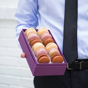 A man with a black tie is holding a box full of French macarons in one hand