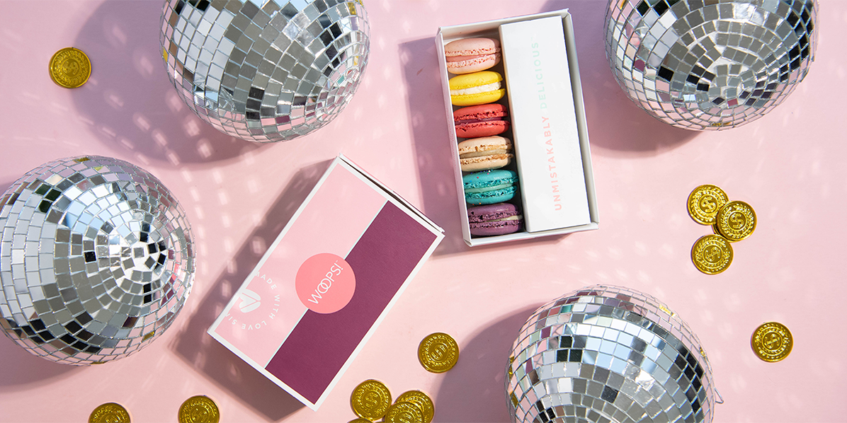 A box full of French macarons and a Woops! Macaron boxes are surrounded by disco balls and golden coins