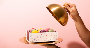  A golden plate with a box full of macarons on top has a golden tray lid being held by a female hand on top. Gold plate and dome