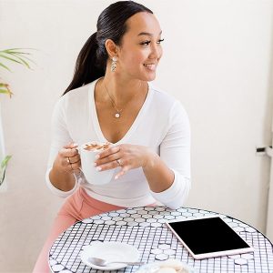A smiling woman holding a coffee has a table with plates full of macarons and an iPad in front of her. 