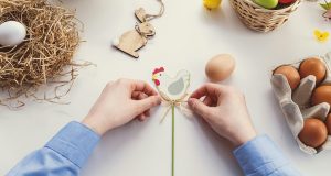 Two hands are tying a ribbon on a DIY hen. Lying around are some Easter eggs, eggs, and some Easter crafts
