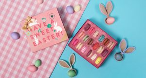 A box full of assorted French macarons has a macaron box with a pink Easter sleeve to the left. Surrounding them are Easter eggs and bunny ears