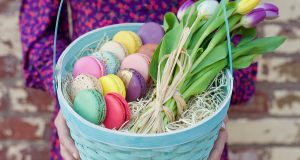 A woman with a red and blue dress is holding a basket filled with macarons, hay, and tulips.