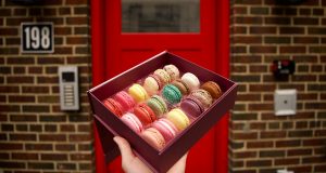 A hand is holding a box full of assorted French macarons in front of a deep red door
