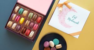 A box full of assorted French macarons has a macaron box with a light blue Eid sleeve and a plate full of macarons to the right