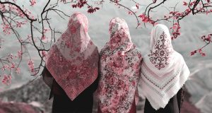 Three women with flowered hijabs are surrounded by pink flowers hanging from a tree.