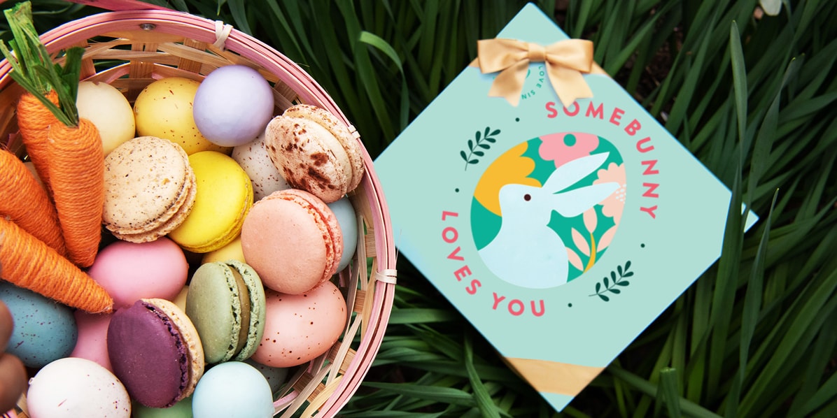 A basket filled with French macarons, Easter eggs, and carrots has a macaron box with an Easter sleeve to the right.
