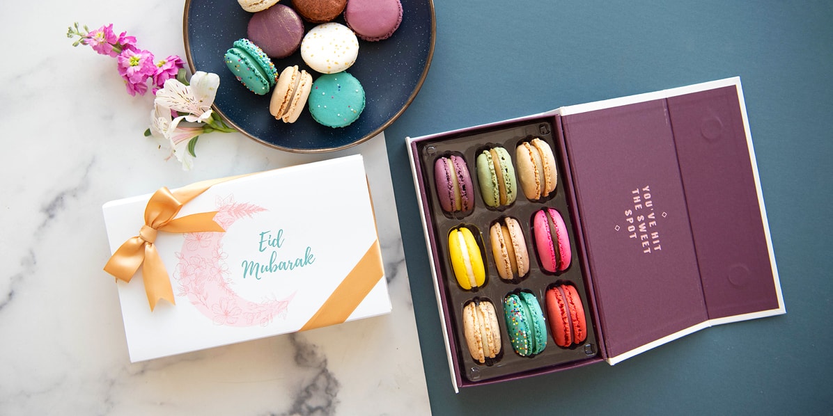 A box full of nine assorted macarons has a macaron box with an Eid sleeve and a plate full of macarons to the right.