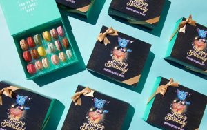 A box full of French macarons is surrounded by macaron boxes with Bingo Blitz birthday sleeves.