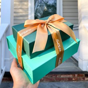 A hand is holding a stack of two green Woops! French macaron boxes tied together by a golden ribbon. The box is in front of a white door.