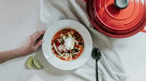 A plate full of food, a white towel, a lemon, a red casserole, and a female hand.