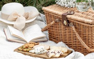 A blanket with a white hat, a book, a picnic basket, and a charcuterie board with cheeses, olives, crackers, and hams. 