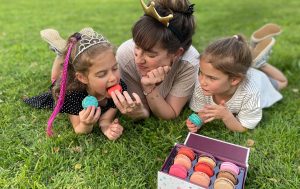 A woman and two girls lying on the grass are eating and holding French macarons in their hands. In front of them is a box full of nine assorted macarons.