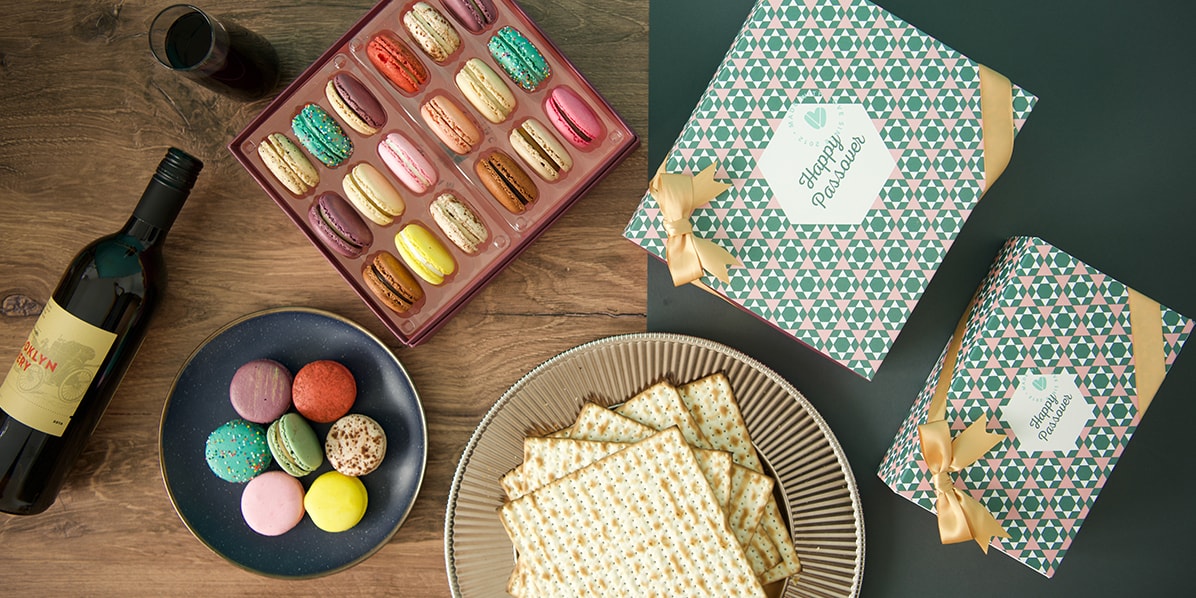 A box full of French macarons is surrounded by macaron boxes with Passover sleeves, a plate full of macarons, another plate full of matzos, and a bottle of wine.