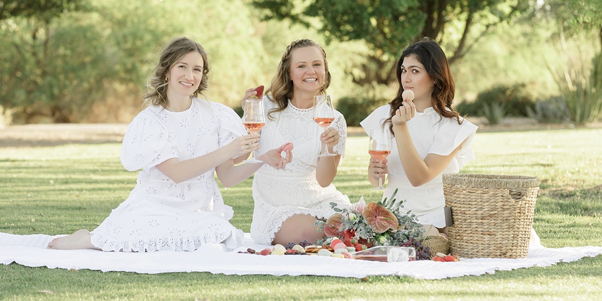: Three women are sitting on a blanket full of the best picnic foods and a picnic basket. They’re holding cups of wine and macarons in their hands.