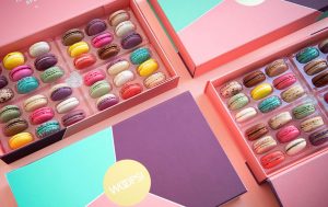  A Woops! French macaron box is surrounded by two boxes full of assorted French macarons.