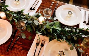 Wooden table with plates, cutlery, tiny candles, glasses, serviettes, and a leaf decoration in the middle.