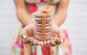  A woman with a flowered dress is holding a French macaron wedding favor box with three French macarons and a golden ribbon in her hands.