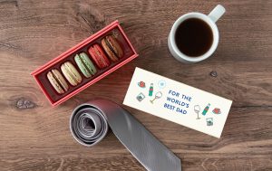 A box full of five assorted macarons surrounded by a macaron box, a cup full of coffee, and a tie. For All Fathers Box of 5