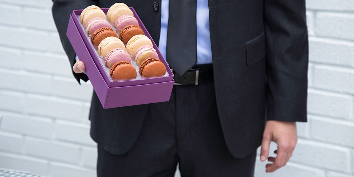 A man in a suit is holding a box full of nine assorted French macarons.