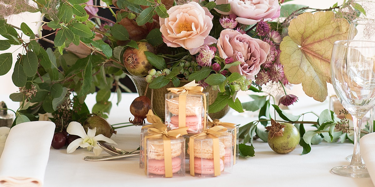 Three French macaron wedding favor boxes with golden ribbons surrounded by glasses, serviettes, and a floral arrangement.