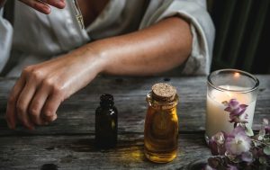 A woman is applying oil to her arm. In front of her is a candle, lavender sticks, and two small bottles of essential oils. 