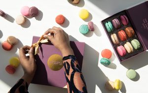 Two female hands are tying a golden ribbon on a purple Woops! Macaron box. Lying around is another box full of macarons and assorted macarons.