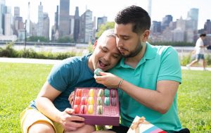 Two men are hugging and sharing macarons. In their laps are Pride Month dessert macaron boxes with Pride sleeves and behind them the New York skyline.