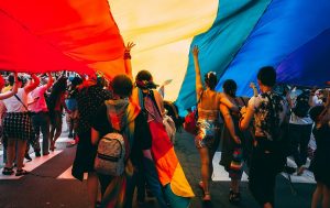 Numerous people dressed in Pride colors walk beneath a big Pride flag.