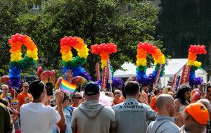 Numerous people with Pride flags wearing colorful clothing and colorful balloons spelling “Pride”.