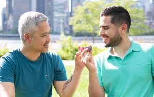 Two smiling men sitting in the grass are holding macarons in their hands. In their laps are Pride Month dessert macaron boxes with Pride sleeves. 