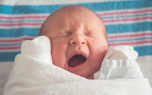 A newborn baby covered in a white blanket is yawning.