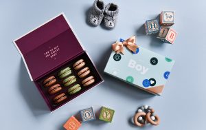 A box full of assorted macarons and a macaron box with a Baby Shower sleeve surrounded by baby toys.