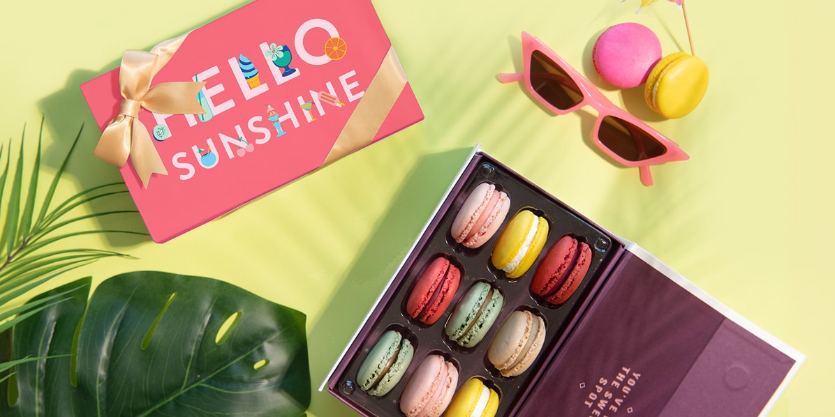 A box full of assorted macarons is surrounded by tropical leaves, a Woops! Box of macarons with a Summer sleeve, a pair of sunglasses, a cocktail umbrella, and two French macarons.