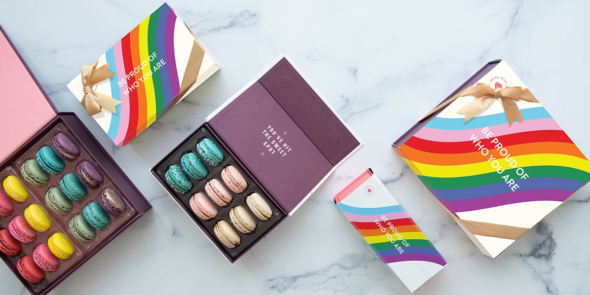 Two boxes full of assorted macarons are surrounded by macaron boxes with Pride sleeves.