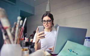 A smiling woman holding her phone and taking a picture. In front of her is a computer and lying around are paintbrushes and papers.