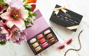 A box full of assorted macarons has a macaron box with a black L’Oreal sleeve, a pair of sunglasses, and flowers on its sides. 