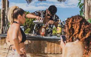 A man preparing a cocktail on tiki-themed cups has two women in front.